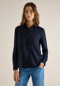 Femme portant une chemise boutonnée bleu marine à manches longues et un jean bleu, debout avec une main dans la poche devant un fond uni.