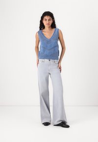 Blauwe gebreide tanktop met een V-hals en chevronpatroon, gecombineerd met lichtgrijze wijde jeans. Zacht materiaal met een ontspannen pasvorm.