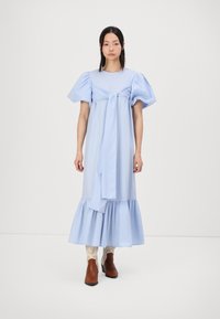 Robe bleu clair, à longueur genou avec des manches bouffantes, cintrée à la taille, dotée d'un détail à nouer et d'un ourlet volanté. Portée avec des bottines marron.