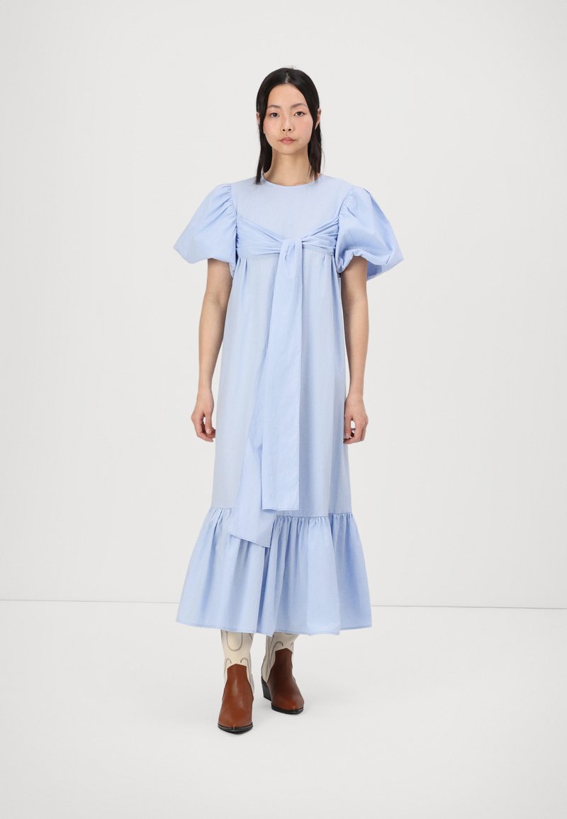 Robe bleu clair, à longueur genou avec des manches bouffantes, cintrée à la taille, dotée d'un détail à nouer et d'un ourlet volanté. Portée avec des bottines marron.