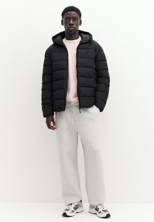 Ung mand iført en sort dunjakke, lyserød sweatshirt, grå joggingbukser og løbesko står mod en ensfarvet hvid baggrund.
