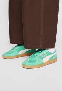 Paire de baskets Puma vert menthe avec semelles en gomme, portées avec des chaussettes blanches et un pantalon large marron.