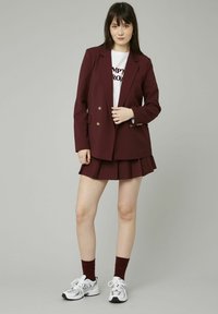 Blazer bourgogne avec des boutons dorés à double boutonnage, associé à une jupe plissée assortie. Porté sur un t-shirt graphique blanc et des baskets blanches.