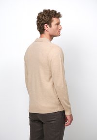 Beige Langarm-Polo-Pullover mit strukturiertem Strick, ausgestattet mit einem gerippten Kragen und Bündchen. Trägt dunkle braune Hosen, seitlich dargestellt.
