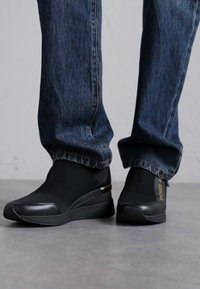 Svart slip-on sko med mesh-overdel, med en glatt gummitåhette og en metallisk detalj på hælen, paret med denimjeans.