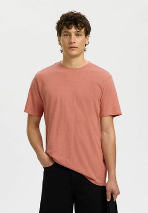 Ung mann med krøllete hår iført en enkel støvete rosa T-skjorte og svarte bukser, stående med den ene hånden i lommen.