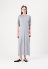 Robe grise, longue, ample et à manches courtes, avec un col rond ; tissu léger avec un léger éclat ; associée à des chaussures plates noires.