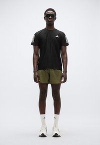 Μαύρο κοντομάνικο πουκάμισο με λευκές ρίγες, πράσινα σορτς, λευκές κάλτσες και αθλητικά παπούτσια. Το μοντέλο φοράει μαύρα γυαλιά ηλίου, στέκεται μπροστά σε λευκό φόντο.