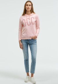 Hellrosa Kapuzenpullover mit langen Ärmeln und bedrucktem Text, kombiniert mit hellblauen Skinny-Jeans mit dezenten Textdetails. Weiße Turnschuhe.
