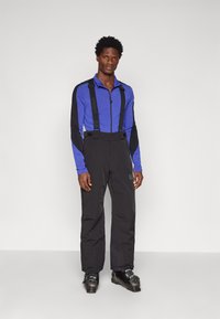 Zils augstā apkakle garām piedurknēm ar melniem akcentiem, kombinēts ar melniem slēpošanas biksēm, kas aprīkotas ar regulējamiem siksnām. Melni zābaki pabeidz izskatu.