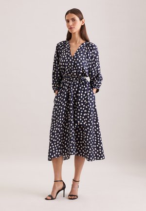 Kvinde iført en midikjole i marineblå med hvide polkaprikker, lange ærmer, V-hals, taljebånd og sorte høje sandaler, stående mod en neutral baggrund.