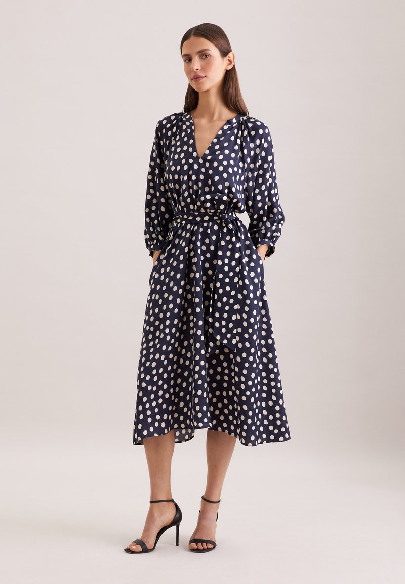 Femme en robe midi bleu marine à pois blancs, manches longues, col en V, ceinture à la taille, et sandales à talons noirs, debout devant un fond neutre.