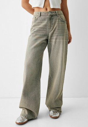 Persoon draagt losse, vervaagde grijze wijde jeans, witte cropped geribbelde top en metallic zilveren sneakers, staande tegen een effen achtergrond.