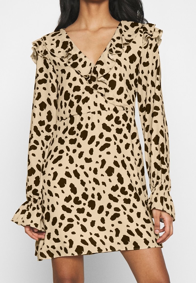 Beige mini-jurk met lange mouwen en donkerbruine luipaardvlekken, met een ruche V-halslijn en gerimpelde manchetten, gedragen door een staand persoon.