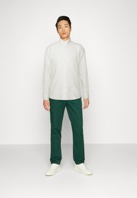 Camisa blanca de botones hecha de un material ligero, combinada con pantalones verde oscuro y zapatillas blancas, de pie frente a un fondo sencillo.