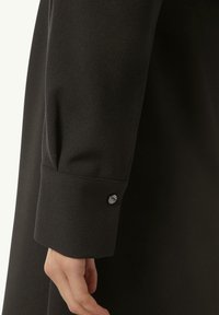 Schwarzer Stoffärmel mit einer glatten Textur, verziert mit einem Knopfdetail nahe dem Bündchen. Das Design ist schlicht und elegant.