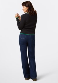 Pull noir à manches longues avec un accent vert sur l'ourlet, associé à un jean évasé bleu. La vue de dos met en avant les détails de broderie sur les épaules.