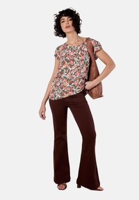 Blusa floreale a maniche corte con motivo rosso, rosa e verde; abbinata a pantaloni svasati marroni; la modella tiene una borsa marrone.