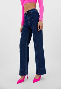 Jeans de mezclilla de tiro alto y pierna ancha en azul oscuro, con costuras amarillas en contraste y cierre de botón, combinados con tacones rosa brillante.
