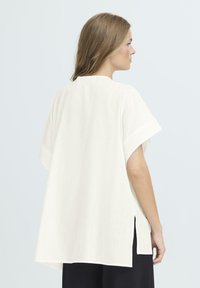 Top bianco, leggero, a maniche corte con fessure laterali, caratterizzato da un tessuto texturizzato e una vestibilità rilassata. Design semplice e minimalista.