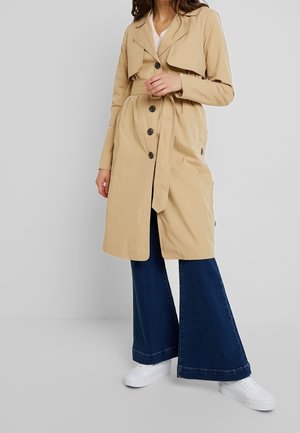 Kobieta ubrana w beżowy płaszcz trencz z paskiem, ciemnoniebieskie dżinsy o szerokich nogawkach i białe trampki na szarym tle.