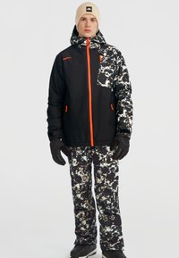 Zwart-wit gestreepte sneeuwjack met oranje accenten en ritszakken, gecombineerd met bijpassende sneeuwbroeken, zwarte handschoenen en een muts.