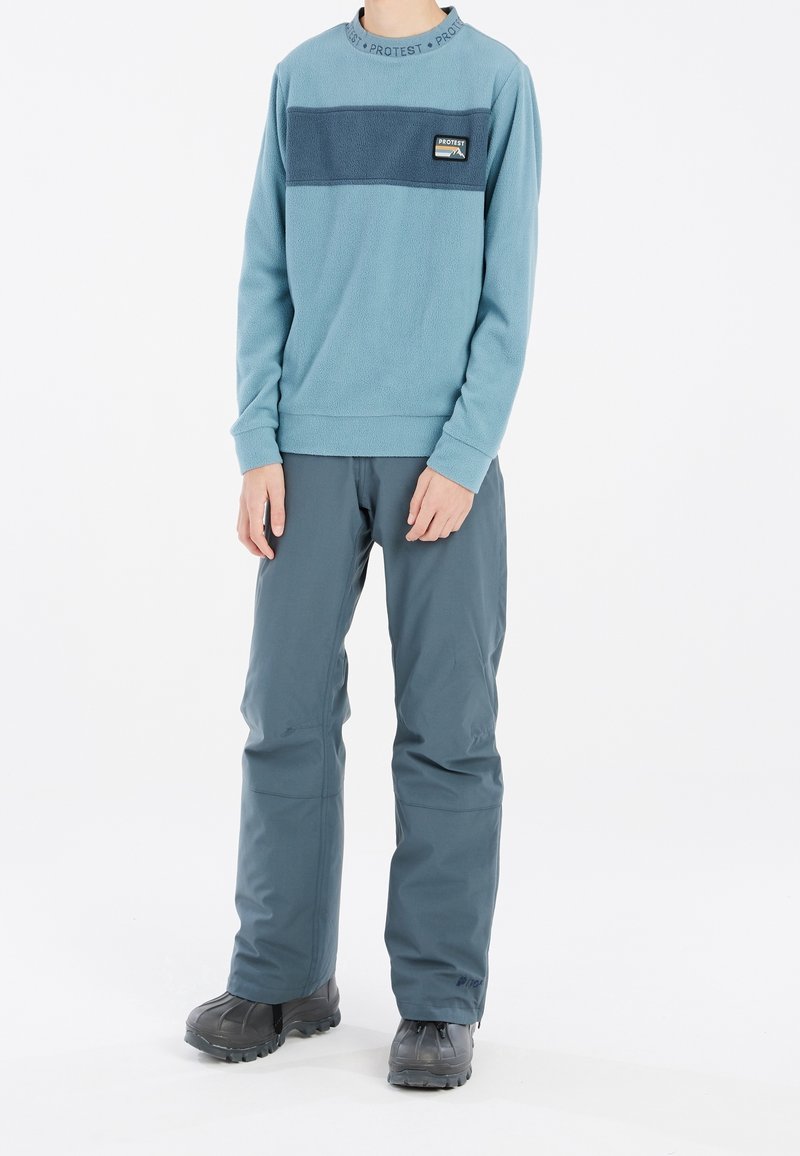 Personne portant un pull en polaire bleu avec une bande bleu foncé et un pantalon gris, debout devant un fond blanc.