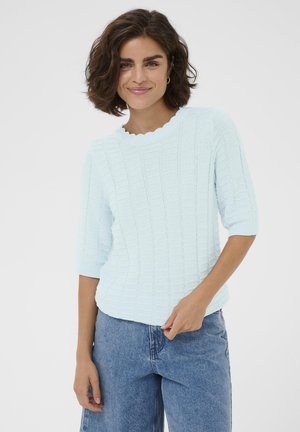 Smiling woman with brown curly hair wearing a light blue textured sweater with scalloped neckline and blue jeans, standing against white background.