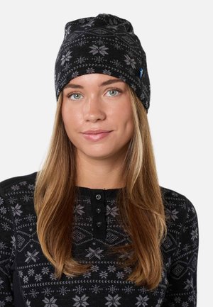 Jeune femme aux longs cheveux châtain clair portant un chapeau noir et un pull avec des motifs flocons de neige gris, souriant légèrement sur un fond uni.
