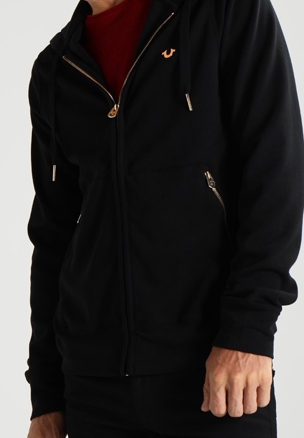 Homme portant un sweat à capuche noir zippé avec des détails dorés sur la fermeture éclair et un petit logo orange sur la poitrine par-dessus une chemise rouge.