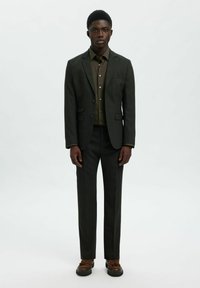 Ciemnozielony garnitur o dopasowanym kroju, z odpowiednią koszulą pod spodem i brązowymi butami. Godnymi uwagi detalami są dwa guziki oraz szyte na miarę spodnie.