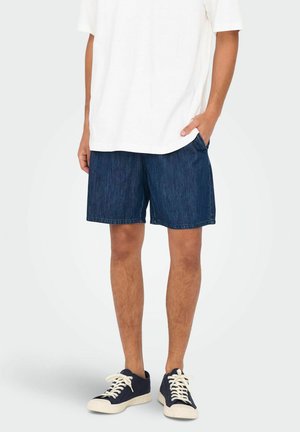 Shorts en denim bleu foncé, en tissu coton, coupe droite, avec poches latérales, portés avec un t-shirt blanc ample et des baskets foncées.