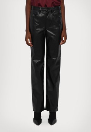 Person trägt hochtaillierte, gerade geschnittene schwarze Lederhose mit Fronttaschen und Knopfverschluss, kombiniert mit schwarzen spitz zulaufenden Stiefeletten.