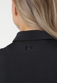 Černé polo tričko s texturovanou látkou, klasickým límečkem a malým lesklým černým logem na zadní straně u límce.