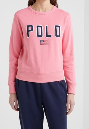 Vrouw draagt een roze sweatshirt met de tekst "POLO" en een patch van de Amerikaanse vlag, gecombineerd met marineblauwe broek.