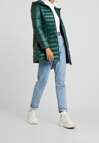 Chaqueta puffy verde con capucha, cierre de cremallera y costuras horizontales. Combinada con jeans azul claro y zapatillas blancas. Silueta ajustada.