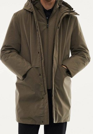 Mann trägt einen langen olivgrünen Wintermantel mit Kapuze, Hände in den Taschen, über einer schwarzen Reißverschlussjacke und schwarzen Hosen.