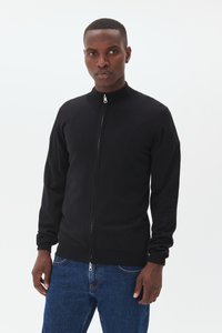 Black zip-up sweater with a high collar, ribbed cuffs, and hem. Knitted texture. Worn with blue jeans. Minimalist design, no visible logos.
