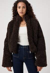 Jachetă scurtă din blană artificială maro cu guler înalt și mâneci wide. Închidere cu fermoar și textură ribbed pe interior, asortată cu blugi albaștri.
