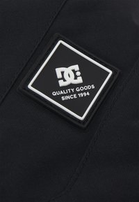 Čierna látková nášivka s bielou embosovanou logom a textom "KVALITNÉ TOVARY OD ROKU 1994" umiestneným v obdĺžnikovom ráme. Textúrovaný povrch.