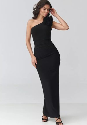 Kobieta w dopasowanej, długiej do ziemi czarnej sukience na jedno ramię z kwiatowym detalem, stojąca z jedną ręką przy włosach, nosząca czarne szpilki.