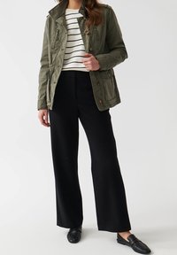 Chaqueta de utilidades verde oliva con botones al frente y bolsillos de parche, usada sobre un suéter a rayas crema y negro, combinada con pantalones de pierna ancha negros y mocasines negros.