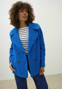 Sininen kaksirivinen takki, jossa on suuret kaulukset, valmistettu teksturoidusta kankaasta. Yhdistettyinä raidallisen paidan ja tummien farkkujen kanssa, malli seisoo yksinkertaista taustaa vasten.