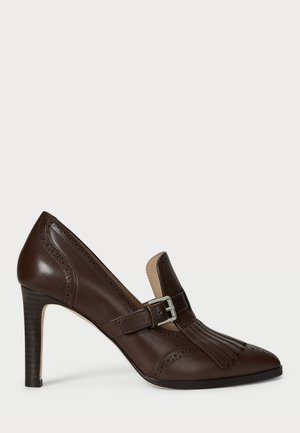 Scarpa con tacco alto in pelle marrone, dalla punta affusolata, con frange decorative e cinturino con fibbia. Presenta dettagli brogue lungo i bordi.