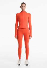 Oranje passende activewear-set met een lange mouwen rits-top en bijpassende leggings, gemaakt van glad, rekbaar materiaal.