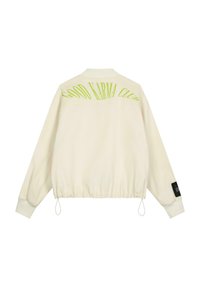 Veste bomber couleur crème avec ourlet et poignets élastiques, arborant le texte vert "GOOD KARMA CLUB" au dos et une étiquette noire sur la manche.