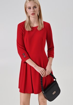 Rode jurk met lange mouwen, geplooide rok en knoopaccenten. Zwarte handtas met gevlochten handvat en gouden logodetail.