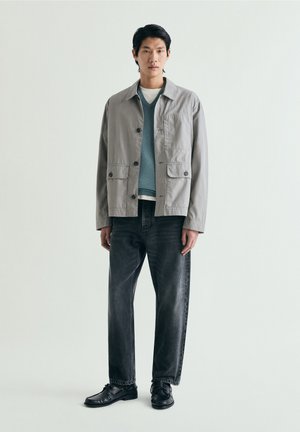 Tânăr stă în picioare purtând o jachetă gri cu nasturi peste un pulover albastru, blugi negri și pantofi negri pe un fundal simplu și deschis la culoare.