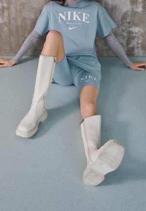 Personne assise par terre portant un short bleu Nike, un t-shirt oversize assorti sur des manches longues grises, et des bottes blanches montantes avec des fermetures éclair.