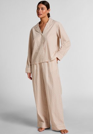 Vrouw staat blootsvoets en draagt een beige-wit geruit pyjamabovenstuk met lange mouwen en bijpassende wijde broek tegen een effen achtergrond.